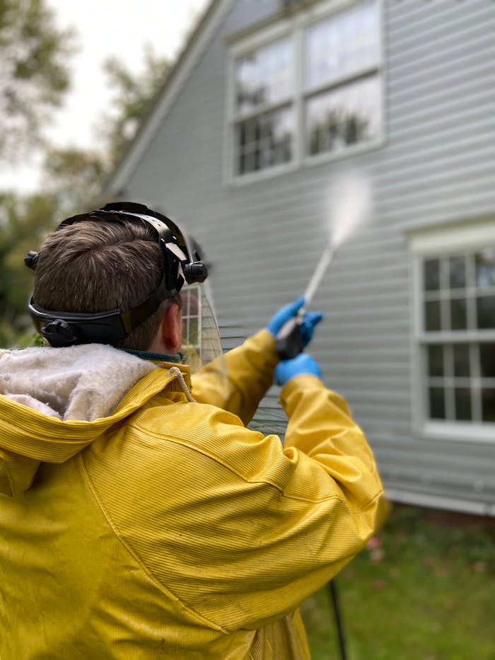 creative Person cleaning house siding with a pressure washer, wearing protective gear in a yellow raincoat.